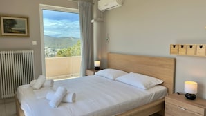 A bedroom with a bed, a nightstand, a lamp, an air conditioning unit, a window with curtains, a radiator, and a wall-mounted shelf. - Delos House Sounio Sea View Temple of Poseidon (Lavreotiki)