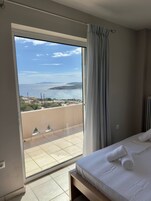 Habitación de hotel con un amplio ventanal corredero que da a un balcón con vistas al mar y colinas al fondo.