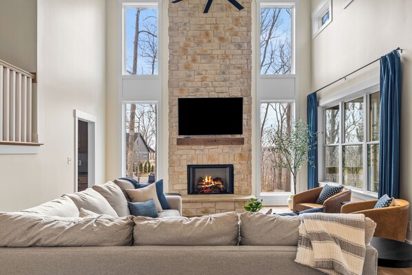 A living room with a stone fireplace, a flat-screen TV, a beige sofa, and a window with curtains.