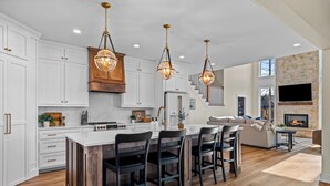 A spacious kitchen with white cabinetry, a large island with bar stools, and a stone fireplace in the living area.