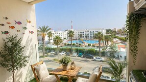 A balcony with a view of a pool and palm trees, a wooden table with chairs, and potted plants. - Appartement S+2 vue mer & golf - Premium hotel residence in Monastir (Monastir)