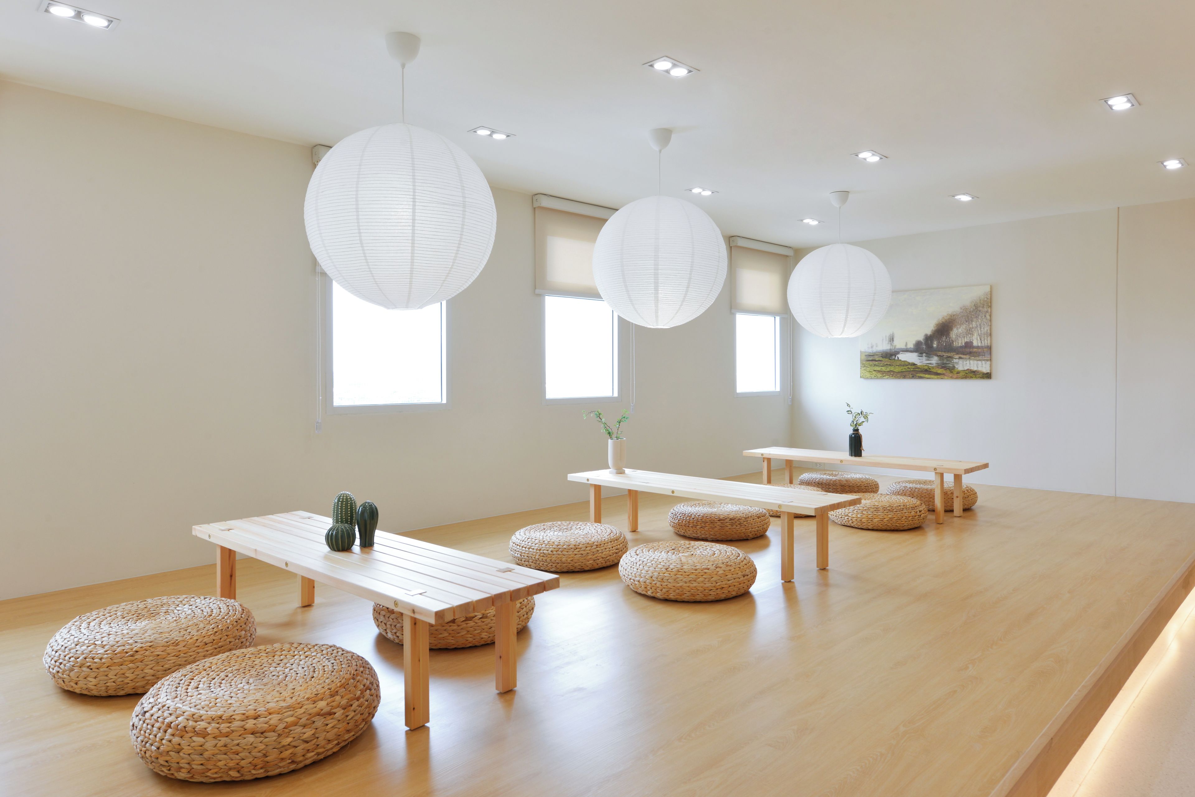 Una habitación minimalista con mesas y bancos de madera, grandes faroles de papel y un cuadro en la pared.