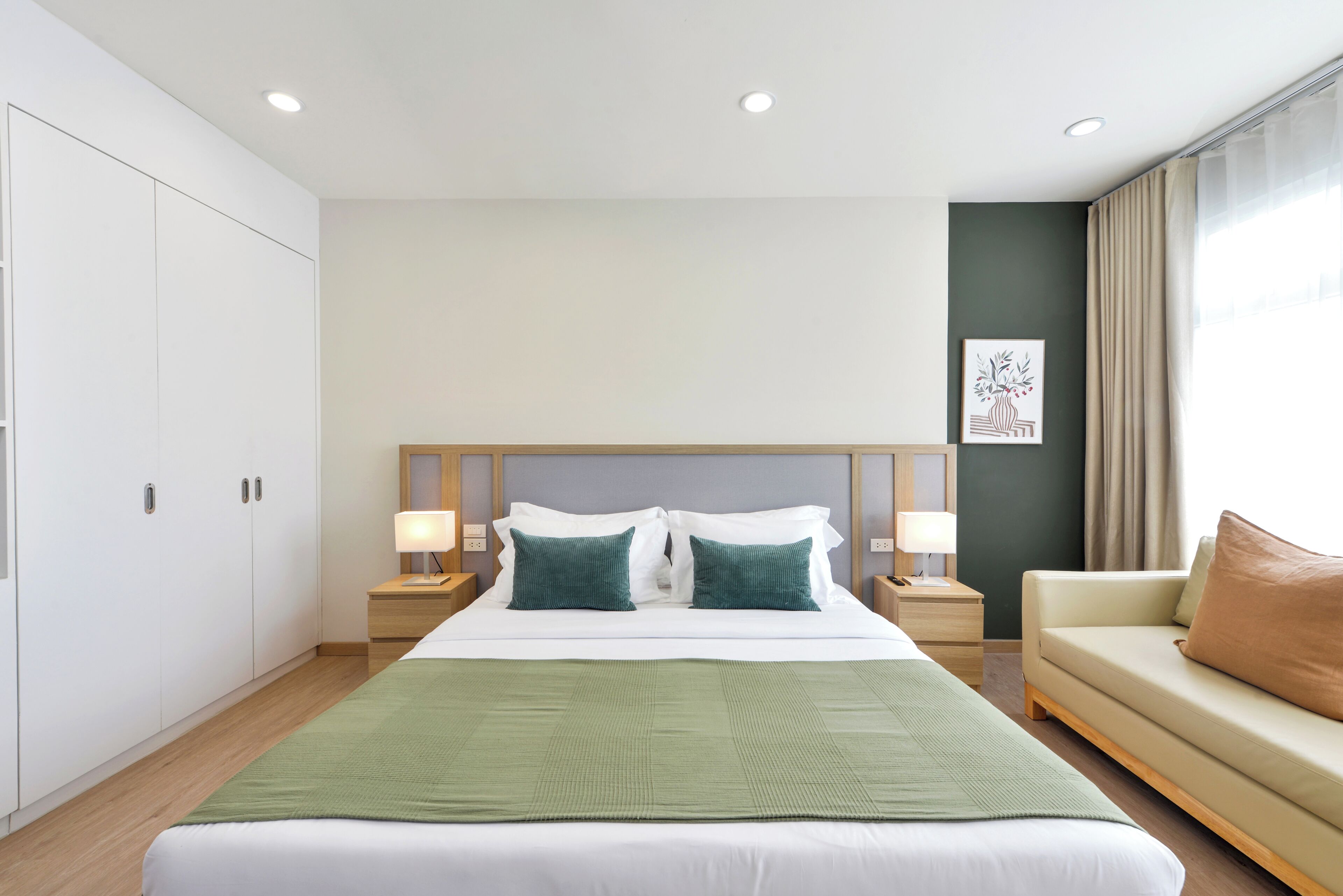 A hotel room with a large bed, two bedside lamps, a green throw, a beige sofa, and a painting on the wall.