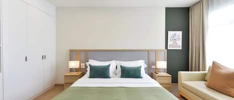 A hotel room with a large bed, two bedside lamps, a green throw, a beige sofa, and a painting on the wall.