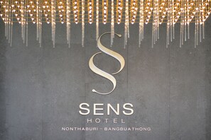Une enseigne d’hôtel avec un grand logo en forme de « S » majuscule et le nom « SENS HOTEL » en dessous, ainsi que l’adresse « NONTHABURI - BANGBUATHONG ».