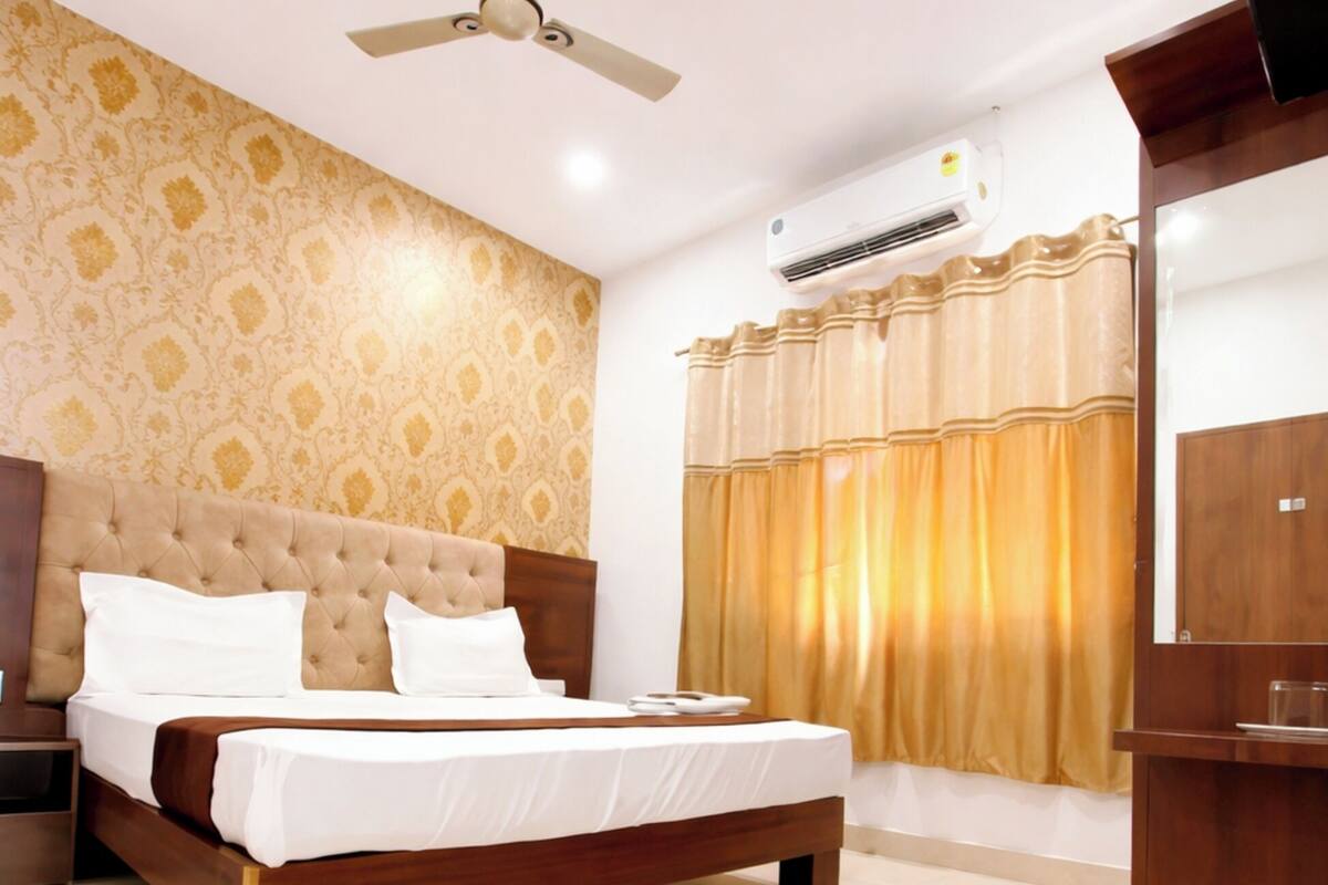 A bedroom with a bed, a ceiling fan, an air conditioning unit, a wooden wardrobe, and a window with curtains.