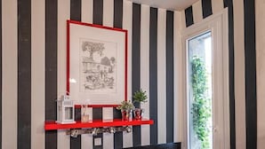 En hall med randiga väggar, en inramad tavla, en röd hylla med dekorativa föremål och en dörr med utsikt över grönskan.