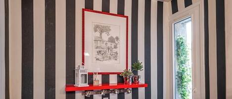 A hallway with striped walls, a framed picture, a red shelf with decorative items, and a door with a view of greenery.