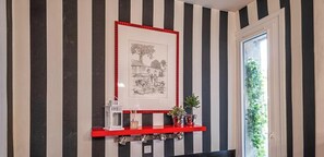 Ein Flur mit gestreiften Wänden, einem gerahmten Bild, einem roten Regal mit Dekoration und einer Tür, durch die Grünpflanzen sichtbar sind.