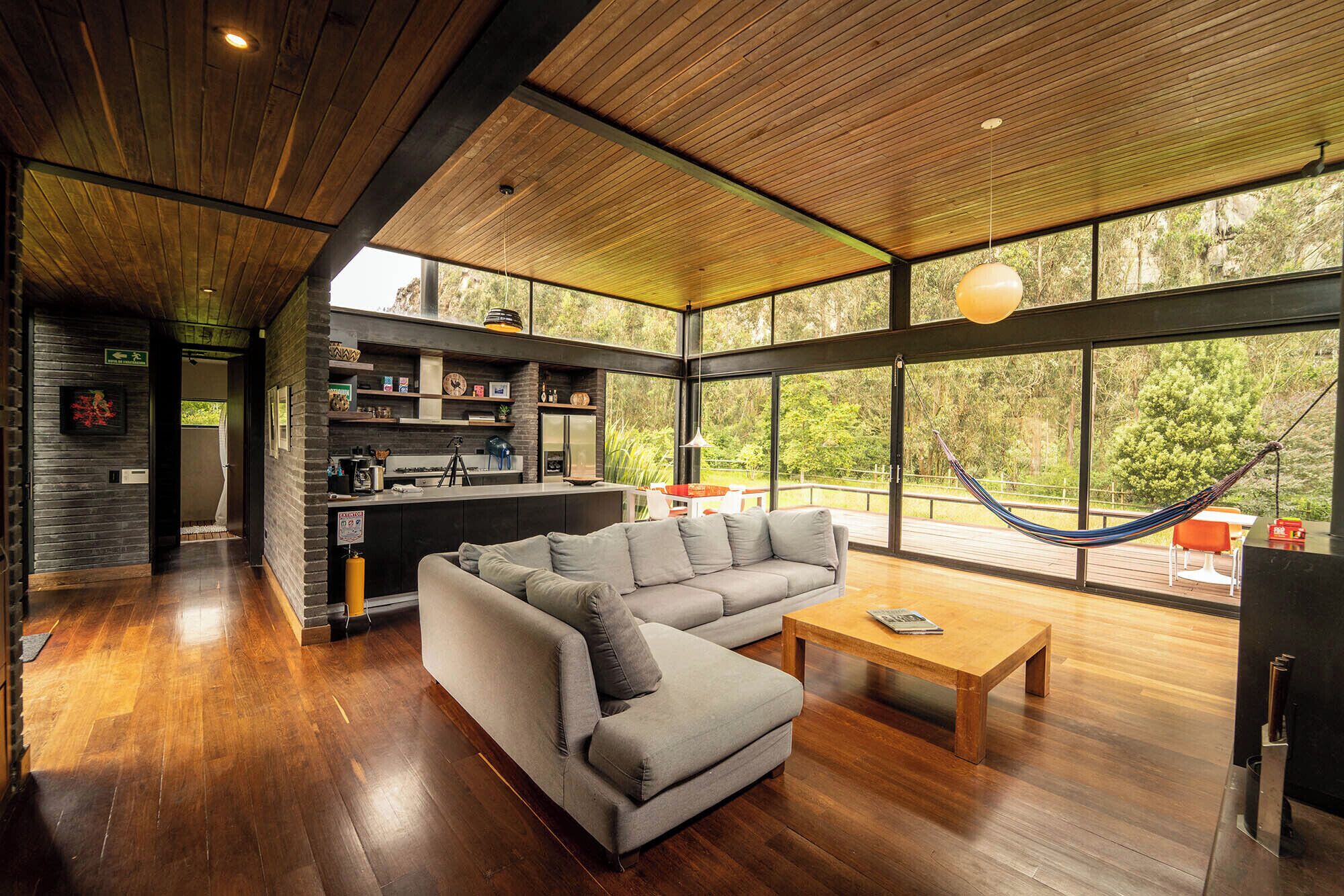 Un intérieur moderne avec un grand canapé d’angle, une table basse en bois et un hamac placé près d’une grande fenêtre donnant sur un espace verdoyant.