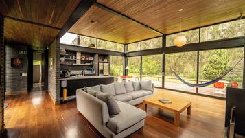Un intérieur moderne avec un grand canapé d’angle, une table basse en bois et un hamac placé près d’une grande fenêtre donnant sur un espace verdoyant.