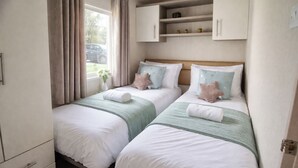 A bedroom with two beds, a window with curtains, a small shelf with decorative items, and a bedside table.