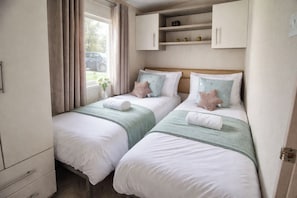 A bedroom with two beds, a window with curtains, a small shelf with decorative items, and a bedside table.