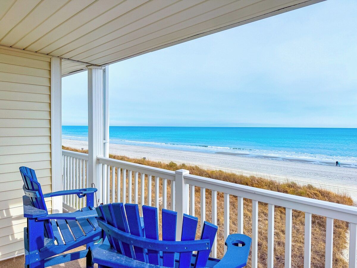 A covered porch with a view of the beach, featuring blue Adirondack chairs.