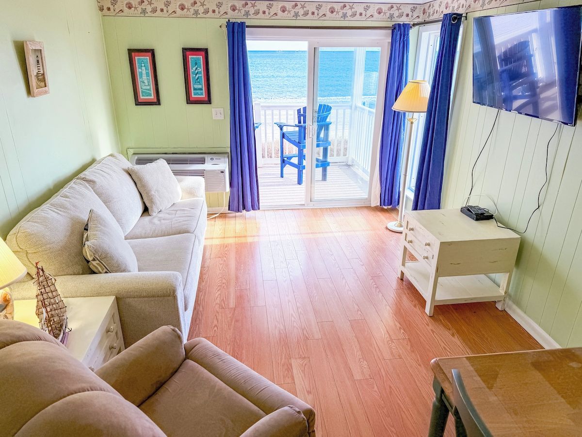 A living room with a sofa, a small table, a desk, and a view of the ocean through a glass door.