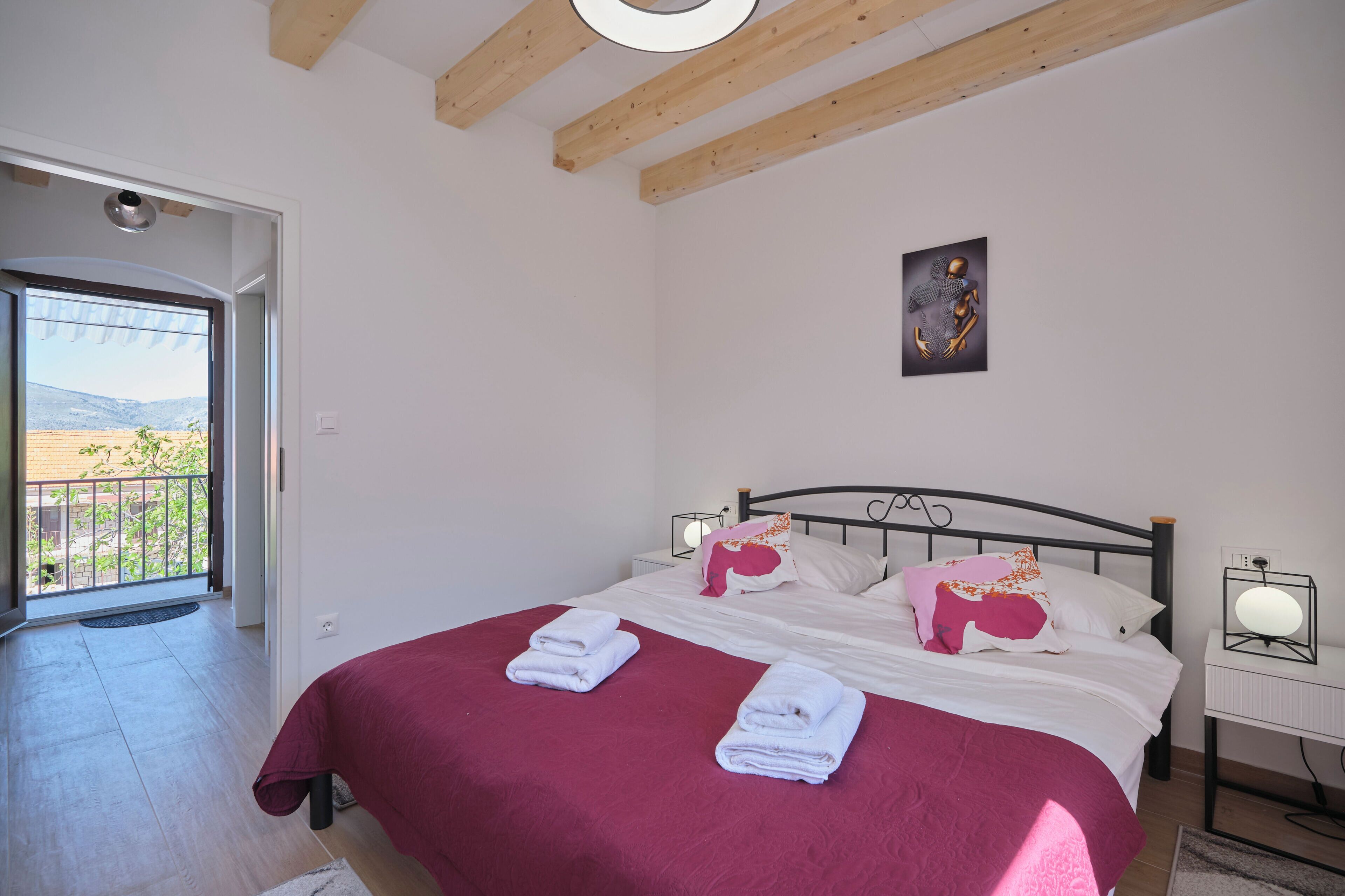 Un dormitorio con cama, almohadas, una mesita de noche con lámpara y vistas a través de una puerta abierta.