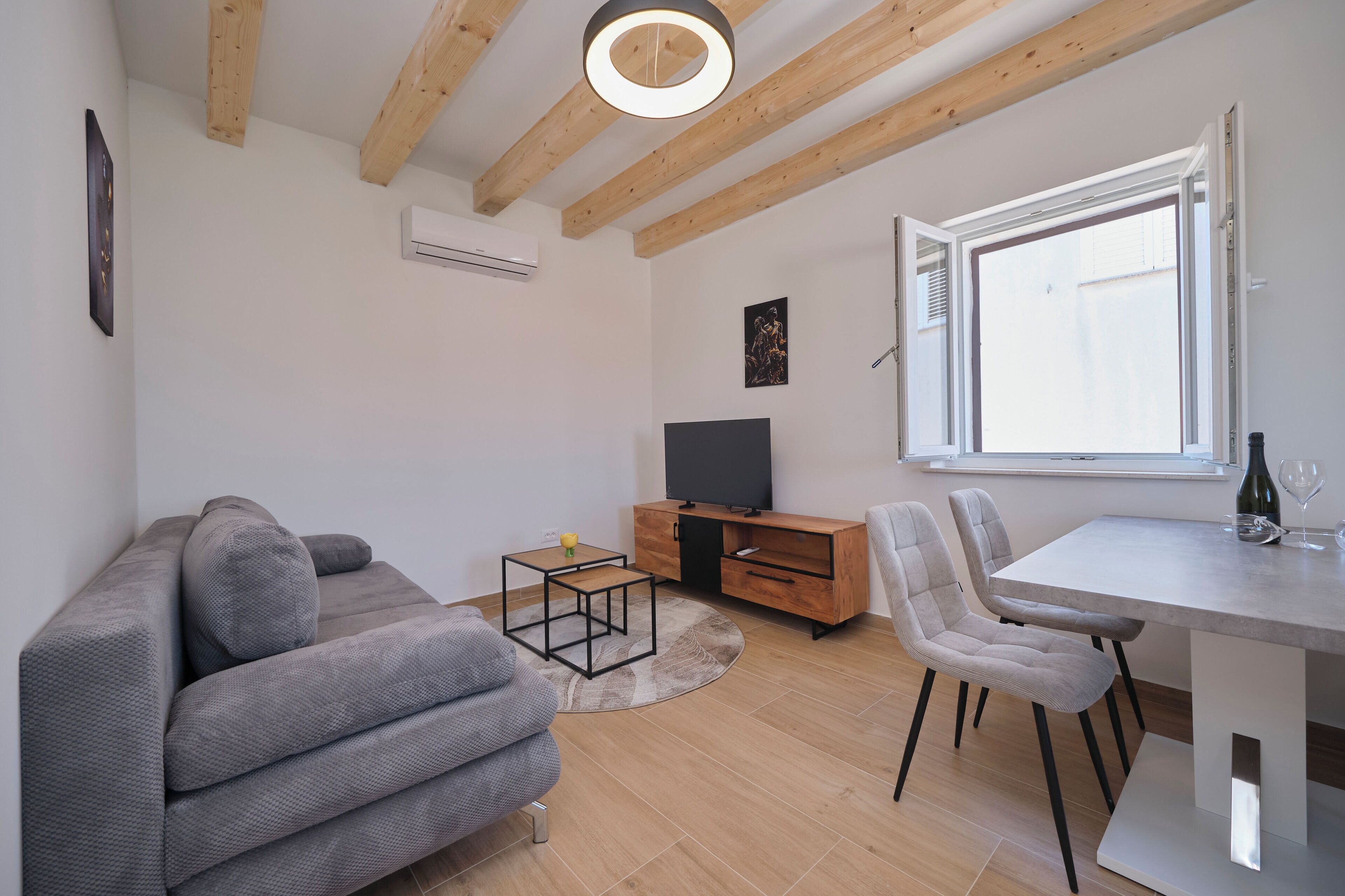 Ein modernes Wohnzimmer mit einer Couch, einem Fernseher, einem Esstisch, Stühlen und einem Fenster mit Jalousien.