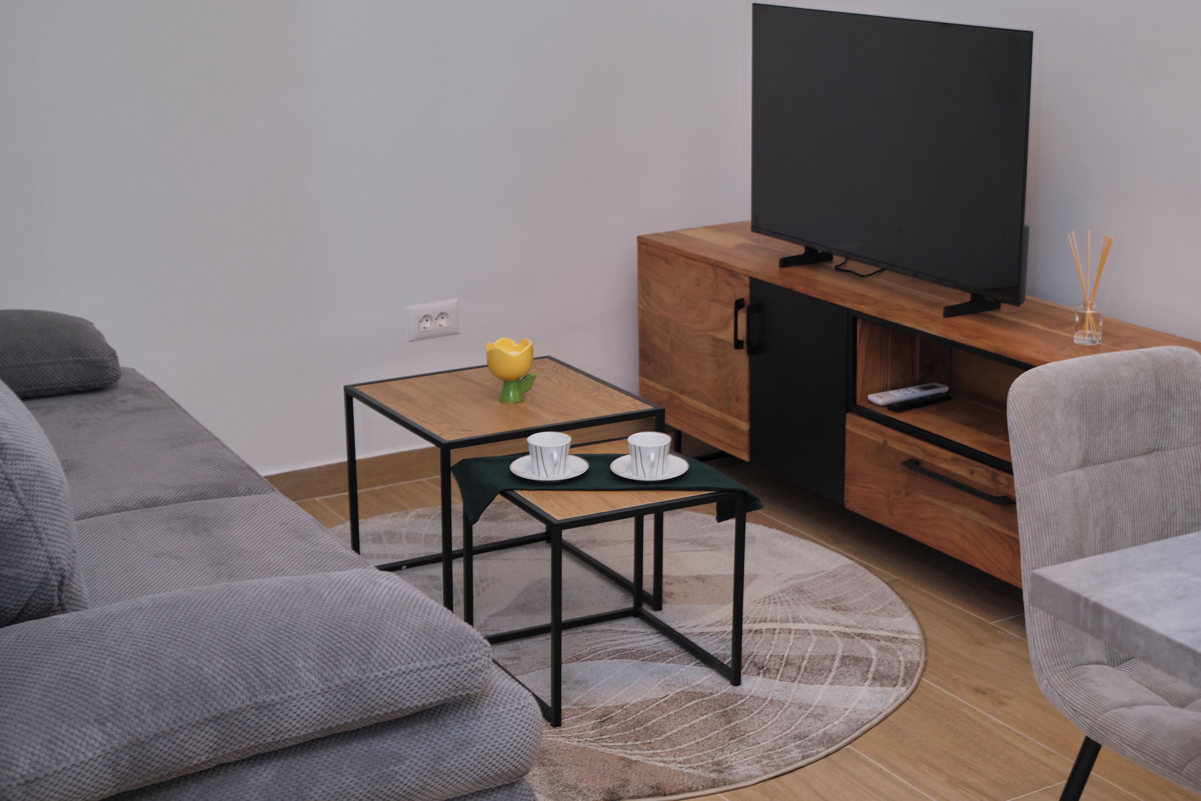 A modern living room with a grey sofa, a small wooden table with two white cups, a yellow flower vase, a flat-screen TV on a wooden cabinet, and a grey armchair.