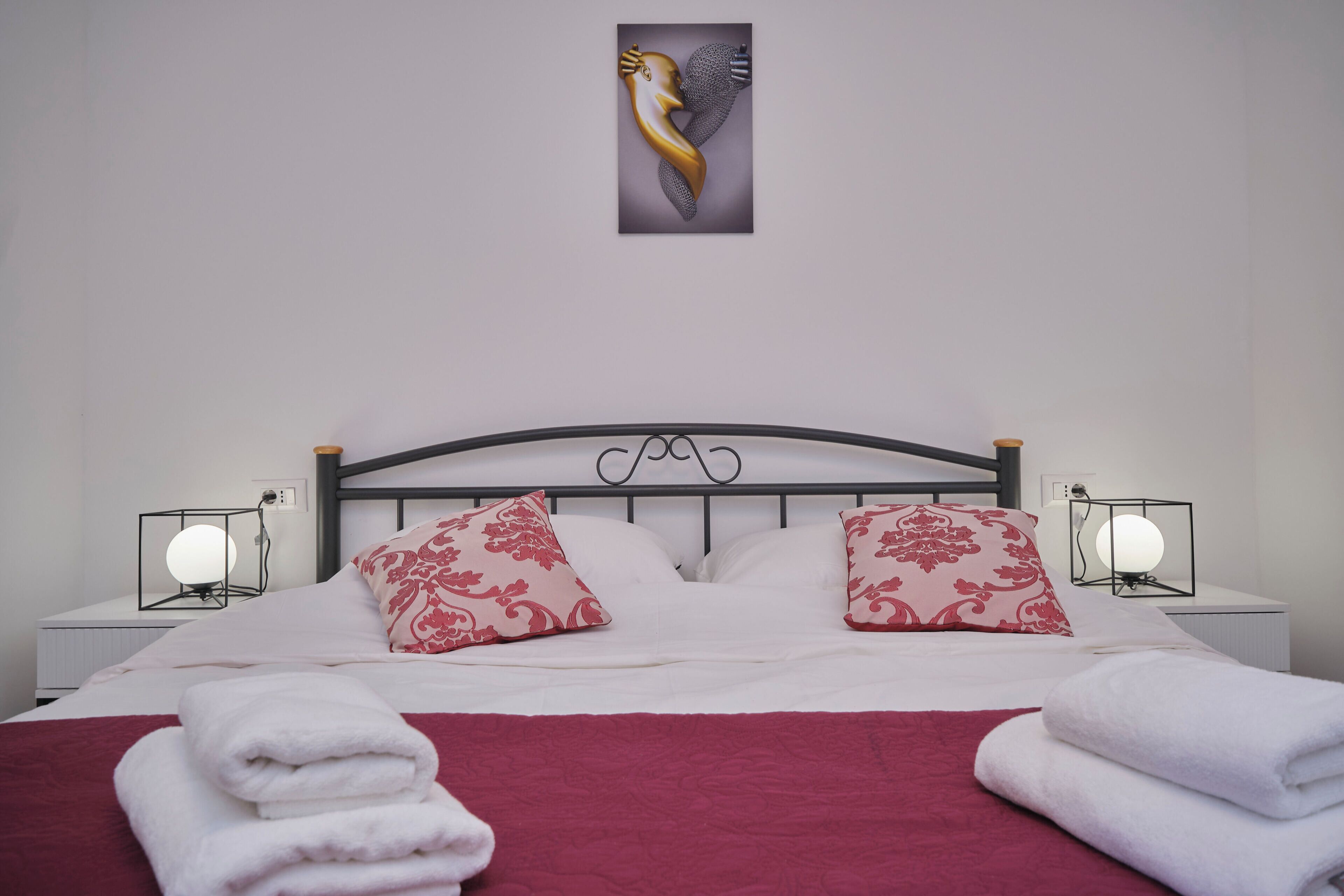 A neatly made bed with white and red bedding, two decorative pillows, and a wall-mounted artwork.