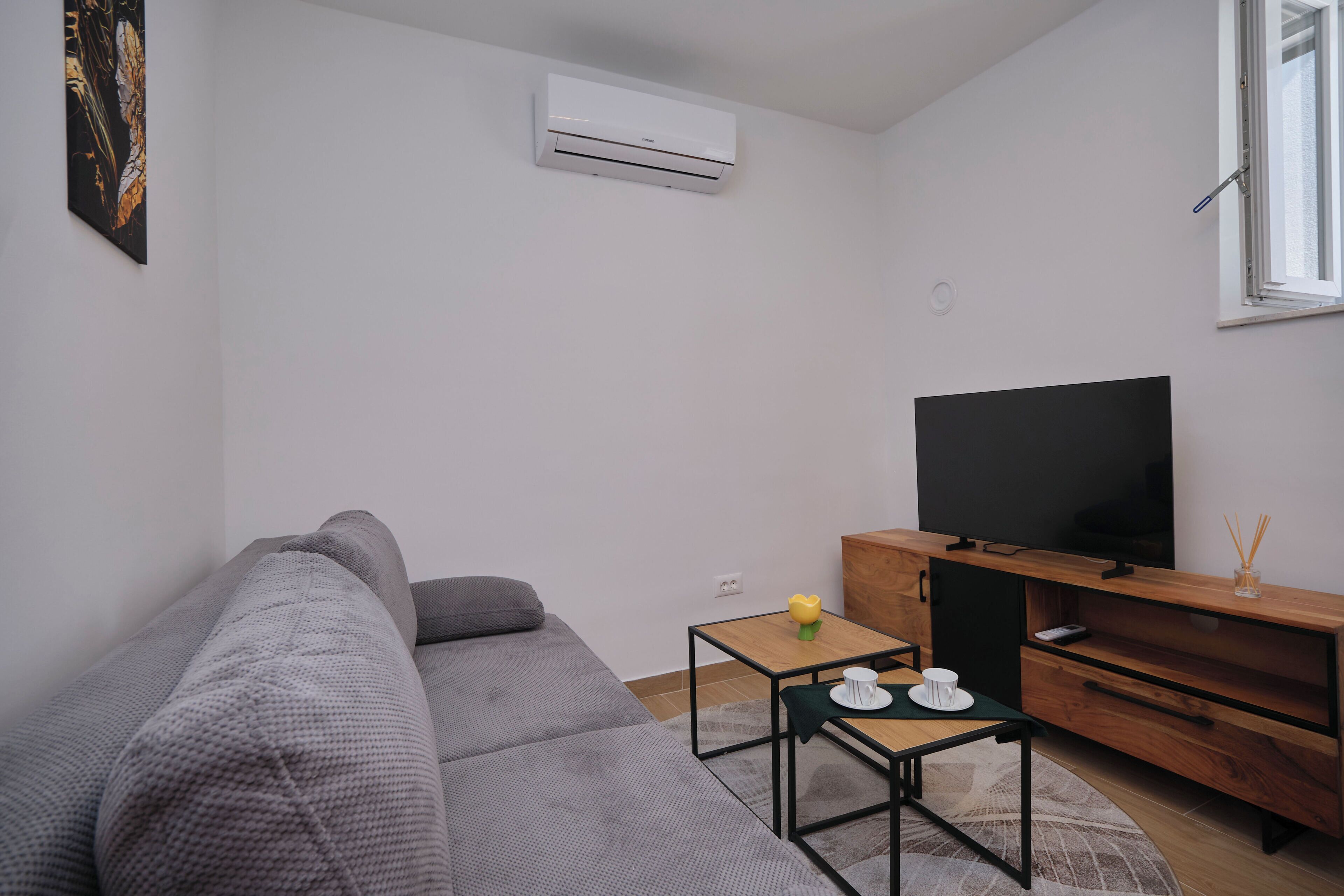 En moderne stue med en grå sofa, et tv-skab i træ, et fladskærms-tv og et lille bord med en kop og en blomst.