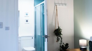 Ein Badezimmer mit Duschabtür aus Glas, einer weißen Toilette, einer hängenden Pflanze, einem Kleiderhaken und einem Nachttisch mit Lampe.