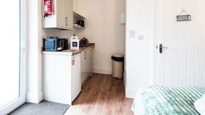 Eine kleine Küche mit Mikrowelle, Toaster und Kaffeemaschine, eine weiße Tür mit dem Schild 'Gone Surfing' und ein Bett mit einer grün gemusterten Decke.