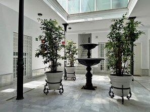 Een ruime binnenruimte met een marmeren vloer, potplanten, een fontein in het midden en een hoge plafond met dakramen.