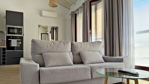 Een moderne woonkamer met een grijze bank, een glazen salontafel en een keuken op de achtergrond.