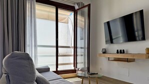 Een moderne woonkamer met een grijze bank, een glazen salontafel, een aan de muur bevestigde televisie en een schuifpui naar een balkon.