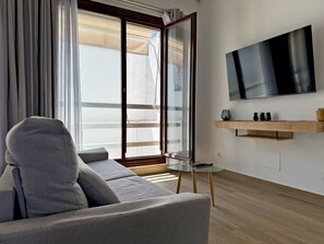 Ein modernes Wohnzimmer mit grauem Sofa, einem Glastisch, einem an der Wand montierten Fernseher und einer Schiebetür zum Balkon.