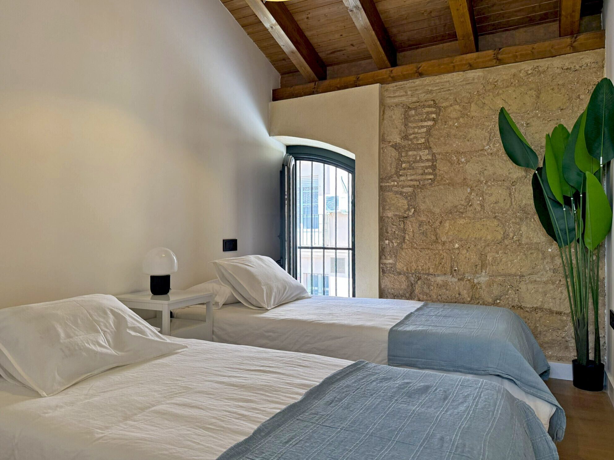 A bedroom with two beds, a small bedside table, a lamp, a large window, and a stone wall.