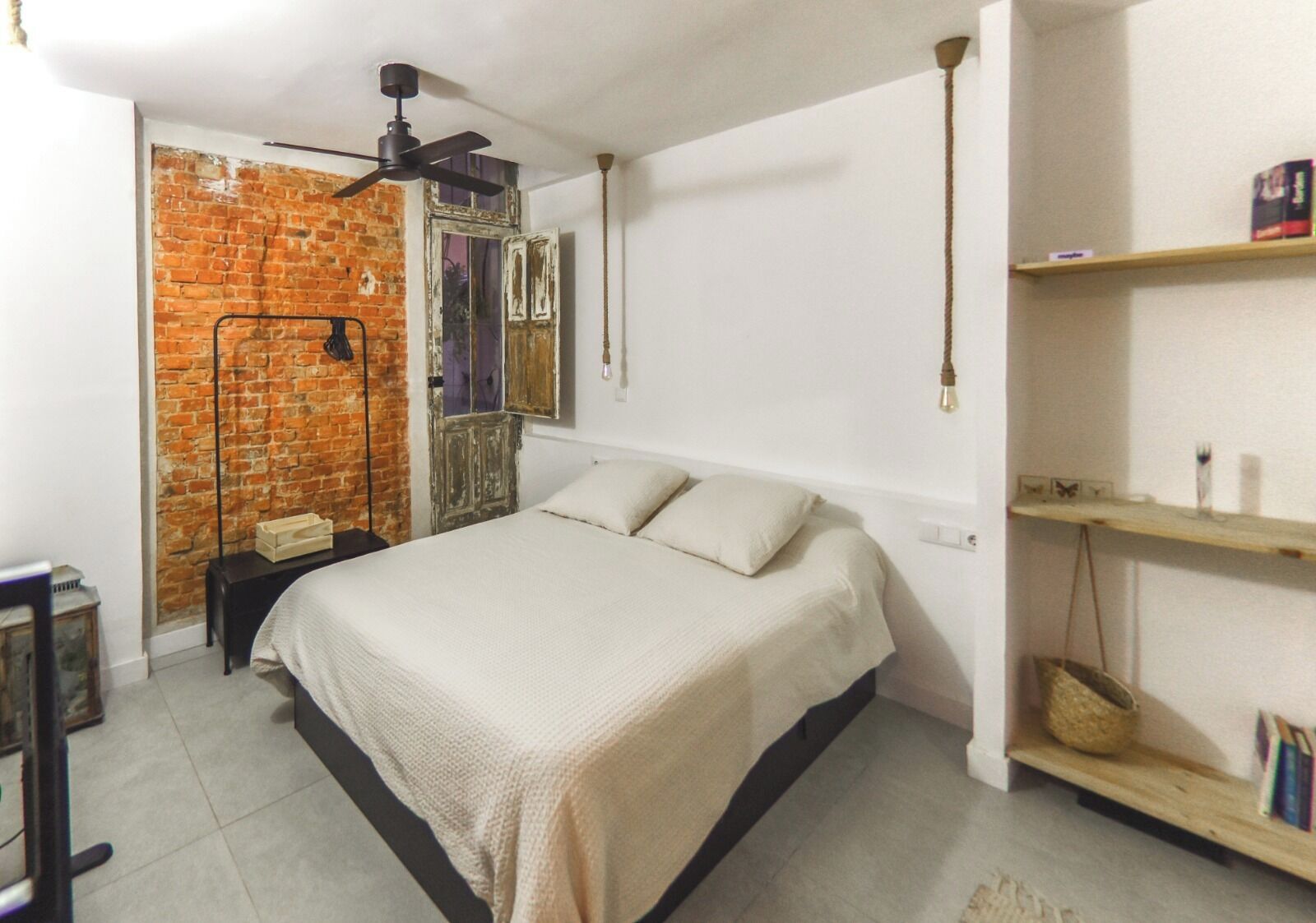 A bedroom with a bed, a brick wall, a ceiling fan, a shelf with books, and a small table.