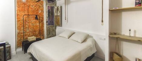 A bedroom with a bed, a brick wall, a ceiling fan, a shelf with books, and a small table.