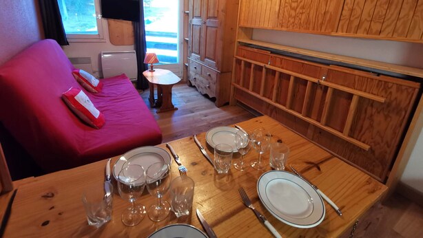 Una habitación compacta con una mesa de comedor de madera preparada para cuatro personas, un sofá rojo con almohadones y una ventana con cortinas.