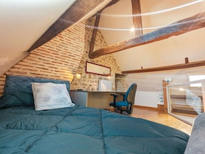 Una camera da letto con soffitto spiovente, un letto con coperta blu, una scrivania con sedia e un muro in pietra.