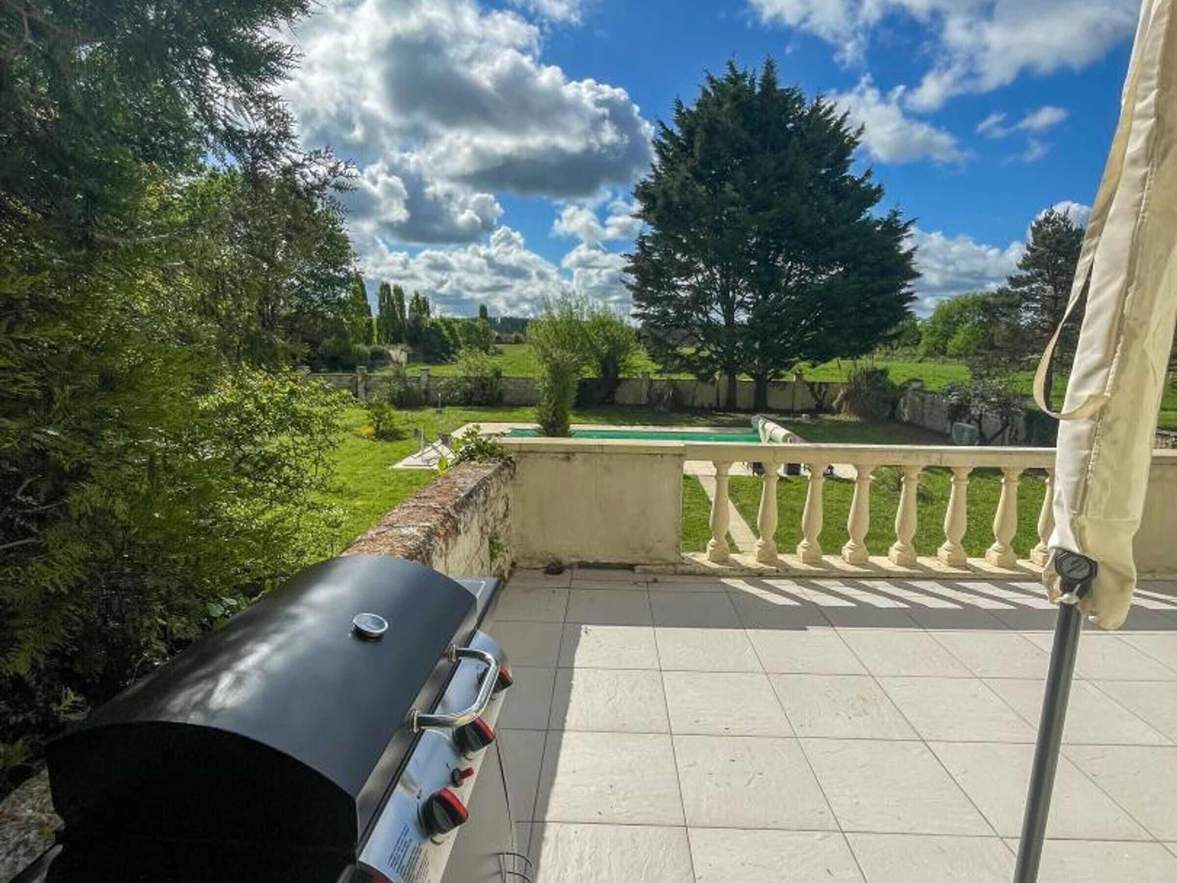 A balcony with a barbecue grill, a view of a green lawn, and a large tree.