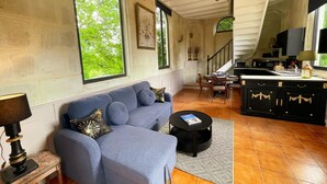 A living room with a blue sofa, a dining area with a table and chairs, a kitchen with cabinets, and large windows with a view of greenery. - Charming chapel, pool near Loire (Bréhémont)
