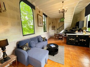 A living room with a blue sofa, a dining area with a table and chairs, a kitchen with cabinets, and large windows with a view of greenery. - Charming chapel, pool near Loire (Bréhémont)