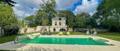 Une piscine avec des chaises longues, un bâtiment en pierre et une végétation luxuriante tout autour.