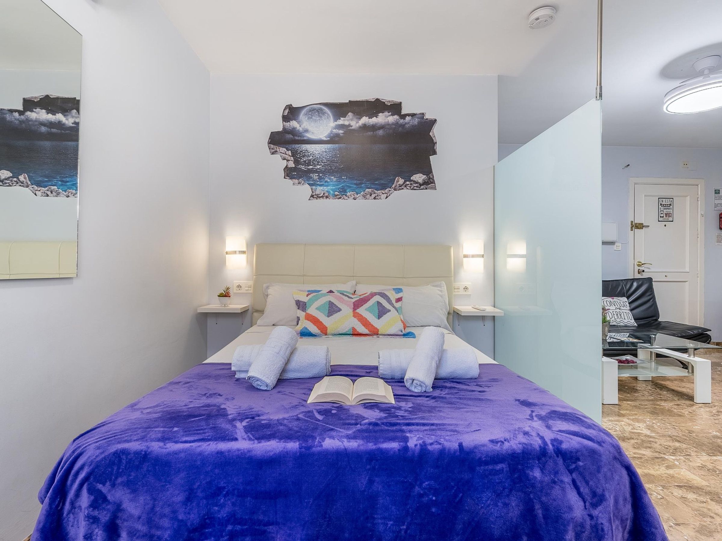 A bedroom with a bed, pillows, a nightstand, a wall art of a moonlit sea, and a door with a sign.