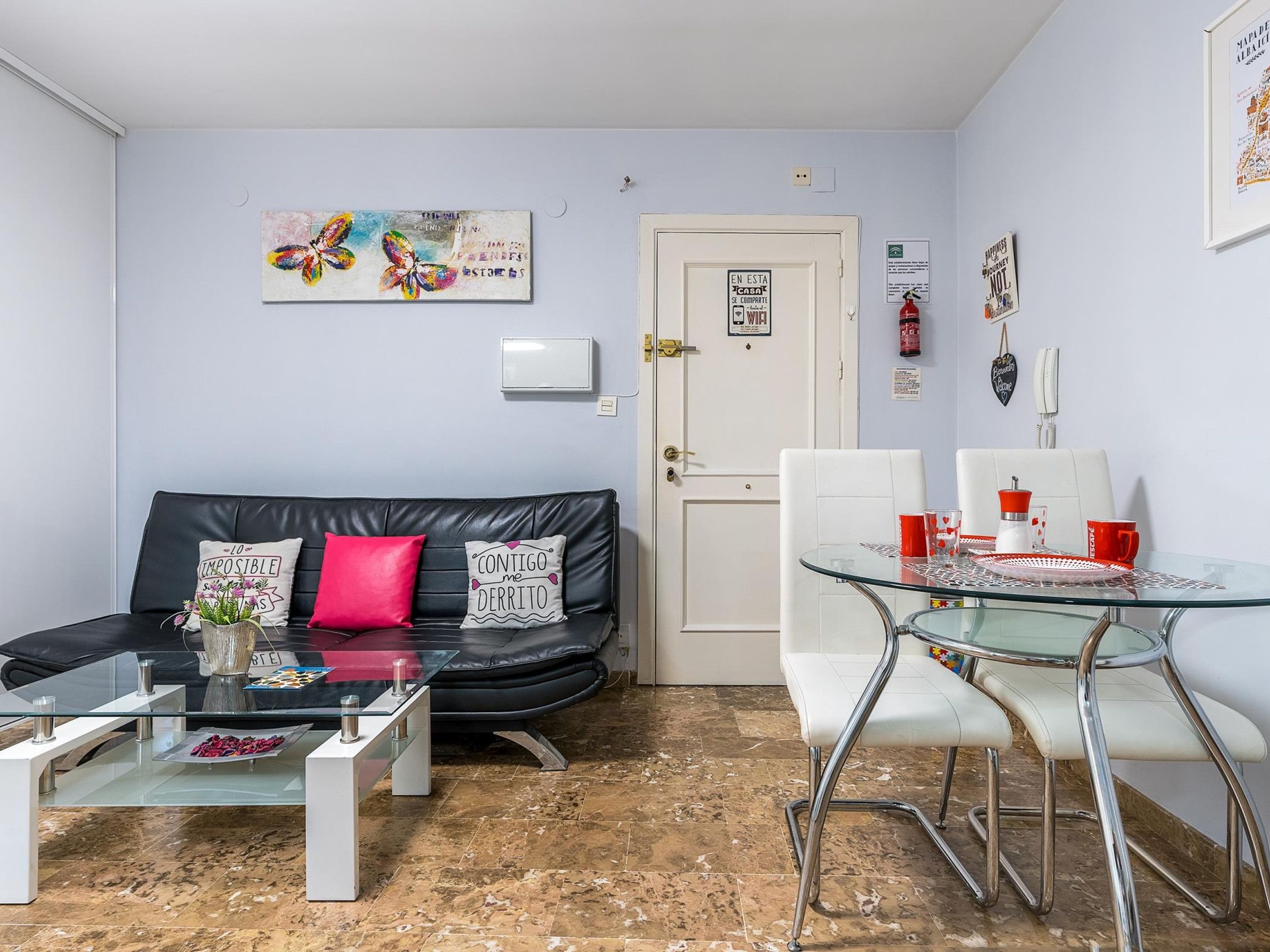 A compact living space with a black leather sofa, a glass coffee table, a white door, and a small dining area with white chairs and red cups.