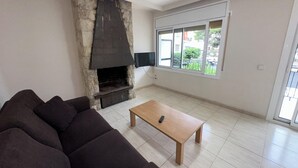 Ein Wohnzimmer mit Kamin aus Stein, einer Couch, einem Holztisch und einem großen Fenster mit Blick ins Grüne.
