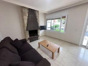 A living room with a stone fireplace, a sofa, a wooden coffee table, and a large window with a view of greenery. - Family apartment in Torredembarra - 4 guests (Torredembarra)