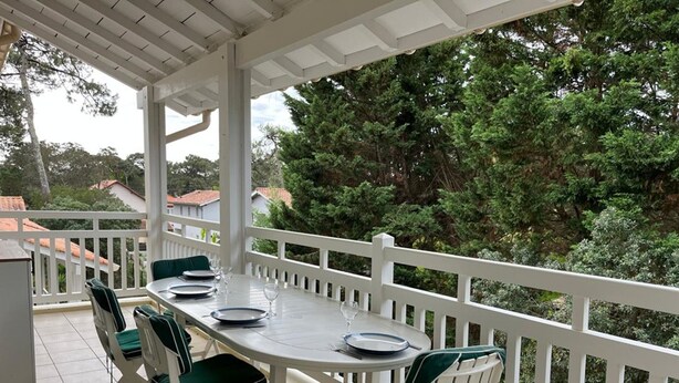 Een overdekt terras met een witte balustrade, een ronde eettafel en groene, beklede stoelen.