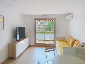 Quarto moderno com televisão sobre um aparador de madeira, varanda com vista, sofá com almofadas amarelas e mesa de jantar.