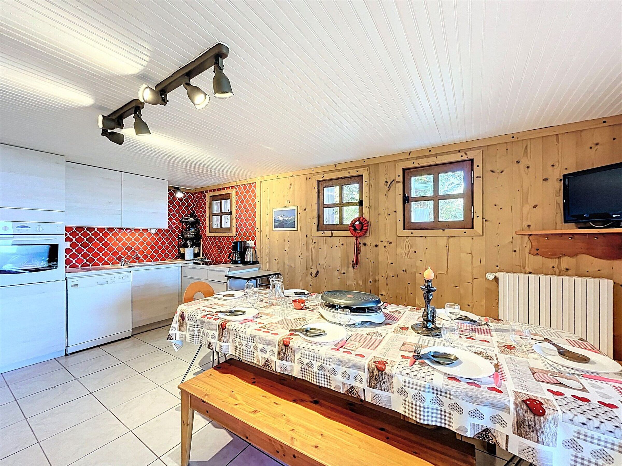 Una cucina con mobili bianchi, un tavolo da pranzo con tovaglia fantasia e un televisore fissato alla parete.