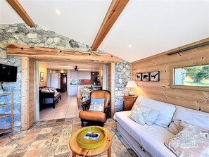 Ein gemütliches Wohnzimmer mit Steinwand, Holzbalken, einer Couch, einem Couchtisch und einem Essbereich mit Tisch und Stühlen.