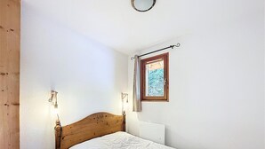 Ein Schlafzimmer mit einem hölzernen Kopfteil, einer weißen Bettdecke, einem Nachttisch, einer Wandlampe und einem Fenster mit Blick auf eine verschneite Landschaft.