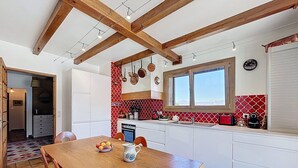 Een keuken met houten balken, witte kasten, een rode en witte achterwand, een eettafel met stoelen en een raam met uitzicht.