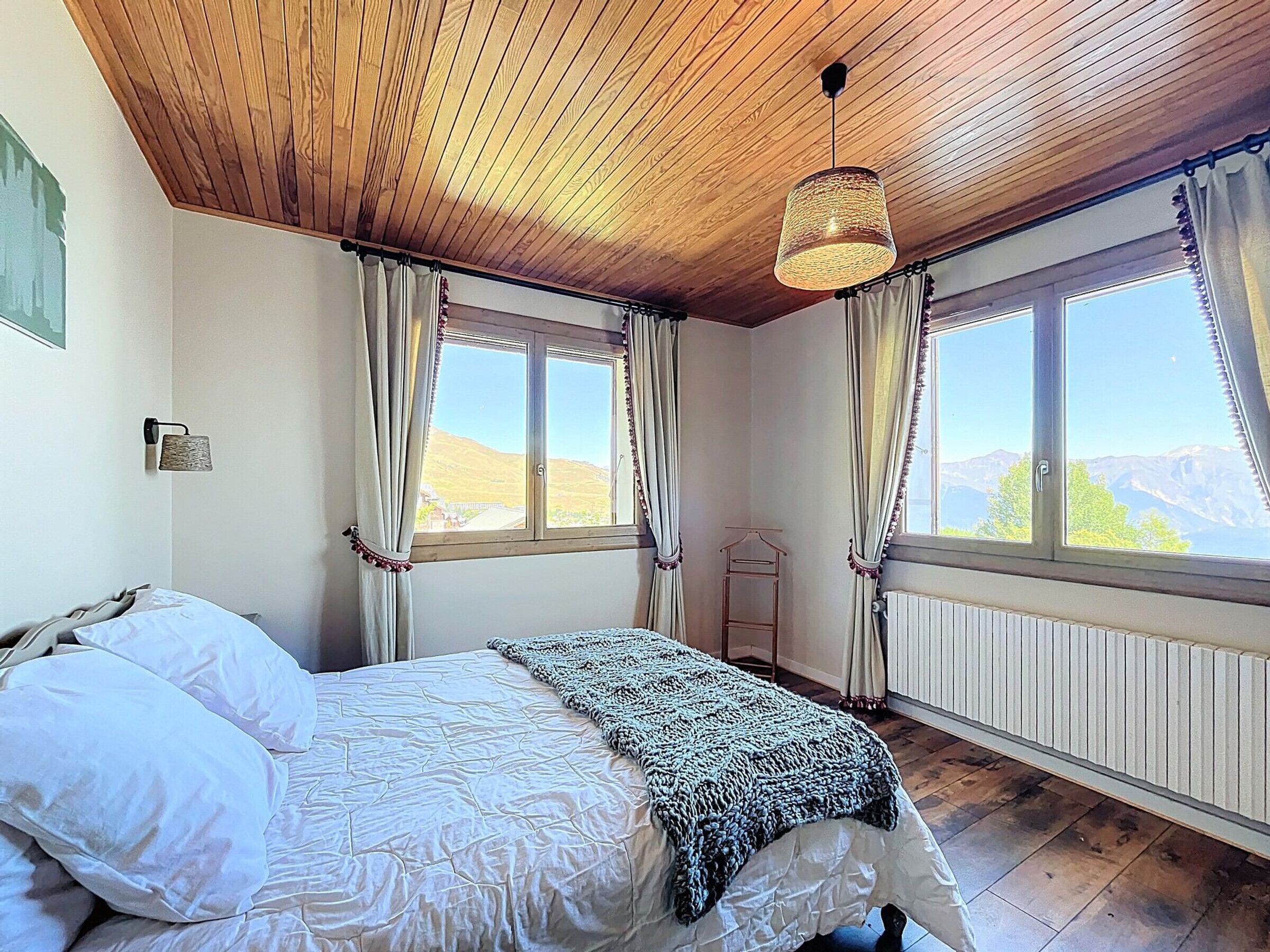 Een slaapkamer met een houten plafond, een bed met wit beddengoed, een gebreide plaid en twee ramen met gordijnen.